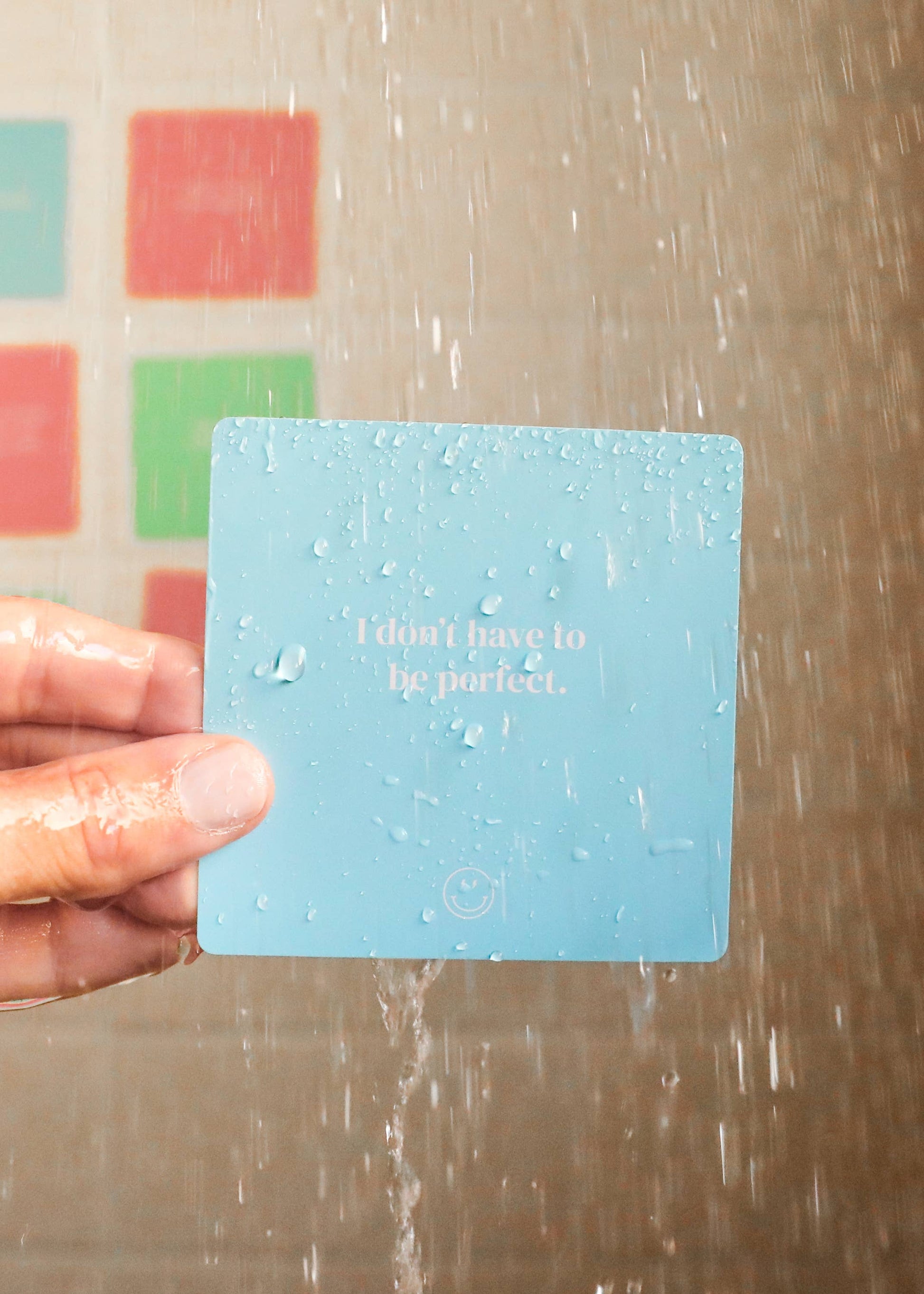 Hand holding a blue card with 'I don't have to be perfect.' text in front of a shower curtain.